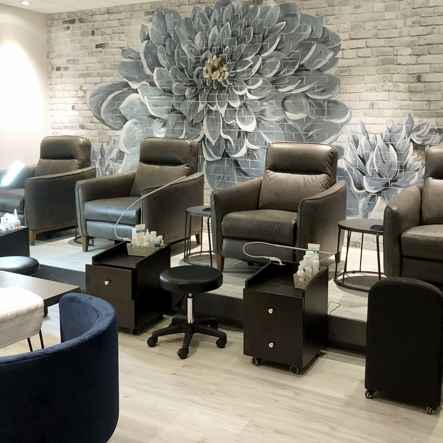 pedicure station at SOHO BARBERSHOP, SPA & HAIR STUDIO, featuring a row of black leather reclining chairs. Brick wall behind has a painting of some giant peonies